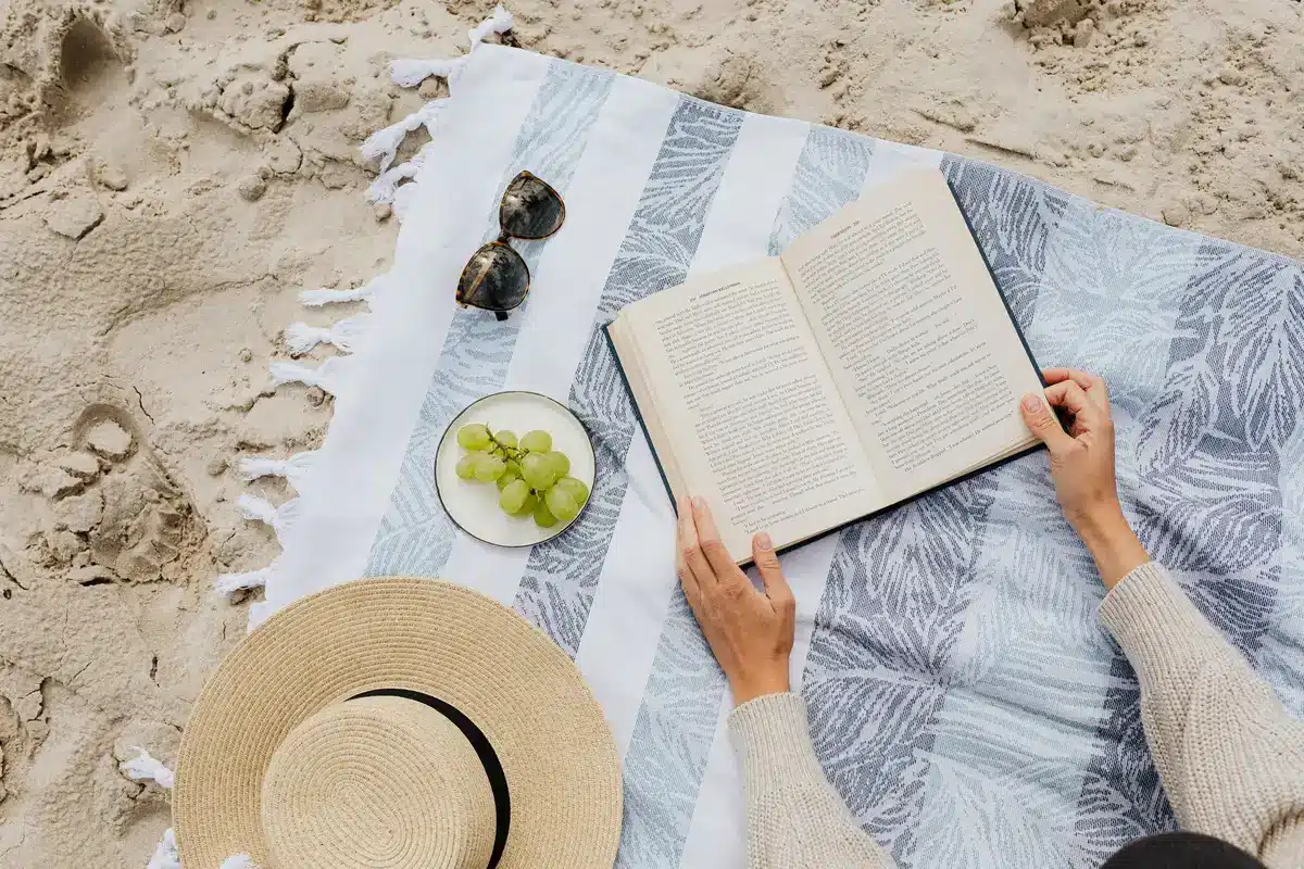 Novelas de romantasy apiladas en una toalla de playa con luz de verano y accesorios de lectura estival.