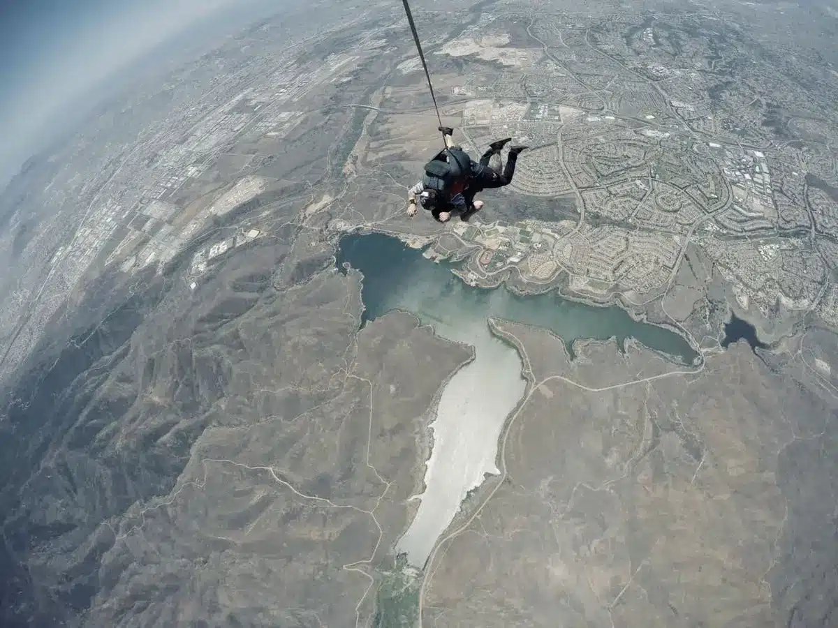 Deportista en caída libre durante un salto extremo, con paracaídas desplegado y paisaje de montañas al fondo.
